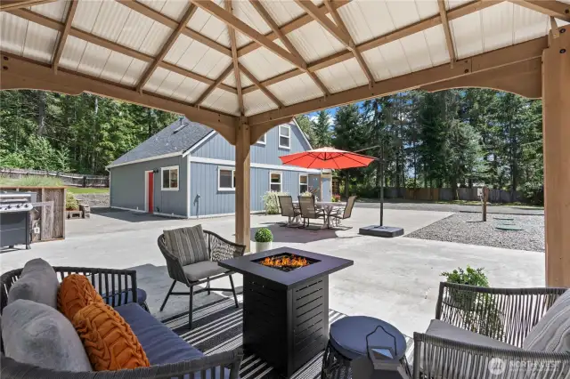 Enjoy seamless indoor-outdoor living with this inviting view from the covered gazebo. The space features a sleek gas fire table, cozy patio seating, and a stylish view of the home's blue exterior and dining patio. The bold red umbrella and grill station add charm and function to this expansive concrete patio, ideal for outdoor dining, relaxing, and entertaining in style.