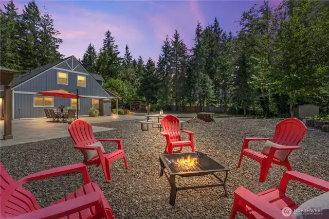 Twilight magic meets modern comfort in this beautifully staged fire pit area with red Adirondack chairs. The soft glow of the fire creates a cozy, welcoming atmosphere with the farmhouse-style home's lights adding warmth in the background. This gravel entertaining area is perfect for late-night conversations, roasting s’mores, or simply relaxing under the stars.