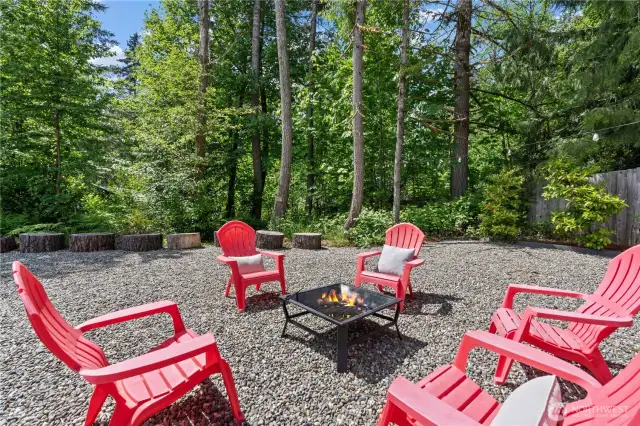 Bright and inviting, this forest-framed fire pit space is ready for unforgettable outdoor moments. Surrounded by mature trees and greenery, the vibrant red Adirondack chairs encircle a square fire feature, offering a perfect place to gather with friends and family. Log stump seats and string lights add rustic charm and functionality to this thoughtfully designed outdoor zone.