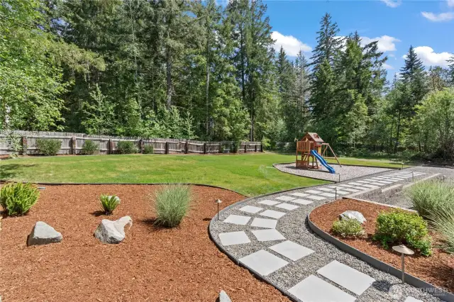Spacious and professionally landscaped backyard featuring a modern paver and gravel walkway, lush green lawn, and private wooded views. Enjoy family time with a built-in playset and bark-covered garden beds with native plants and decorative stones. Fully fenced yard ideal for pets and outdoor entertaining. A Pacific Northwest dream with privacy, play space, and low-maintenance beauty—perfect for families looking for outdoor living.