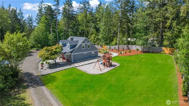 A stunning aerial view reveals the expansive backyard, complete with a fully-fenced grassy lawn, professional landscaping, and a dedicated play area with swings and a slide. The stylish paver pathway leads to the charming modern farmhouse, framed by mature trees and natural privacy. Perfect for families who value outdoor living, safety, and space to roam.