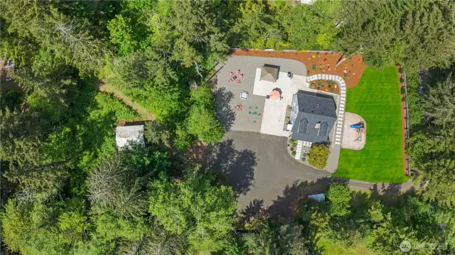 Bird’s-eye view of this beautifully maintained property showcases a large circular driveway, extensive gravel parking, covered patio with outdoor dining area, and fire pit lounge. The vibrant green irrigated lawn, extensive landscaping, and forested perimeter provide a rare combination of function and beauty—ideal for entertaining, homesteading, or simply enjoying the tranquility of your private retreat.