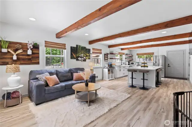 This open-concept living area effortlessly blends cozy charm with modern updates. Featuring rich exposed wood beams, luxury vinyl plank flooring, and a spacious white quartz kitchen island with bar seating, this space is designed for comfort and connection. Natural light floods the room through large windows, while custom open shelving and stainless steel appliances elevate the kitchen’s appeal.