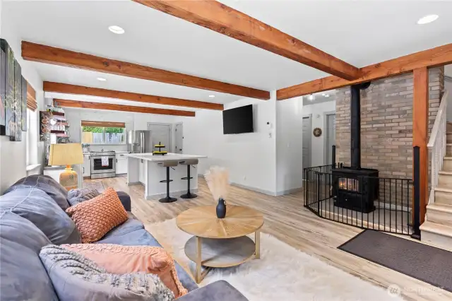 This cozy and functional living area features a charming wood-burning stove with brick surround—perfect for chilly Pacific Northwest nights. Overhead wood beams add warmth and character, while the spacious layout flows directly into a modern kitchen with bar seating. Ideal for entertaining or family nights in.