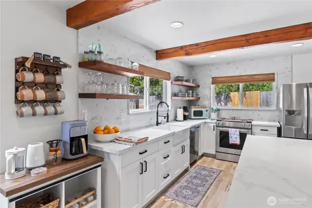 Enjoy modern convenience and rustic charm in this beautifully designed kitchen. Featuring a custom coffee station, a farmhouse-style apron sink, and stunning wood open shelving, this space blends functionality with personality. Natural light pours in through dual windows, accentuating the bright white cabinetry, textured backsplash, and warm wooden ceiling beams.