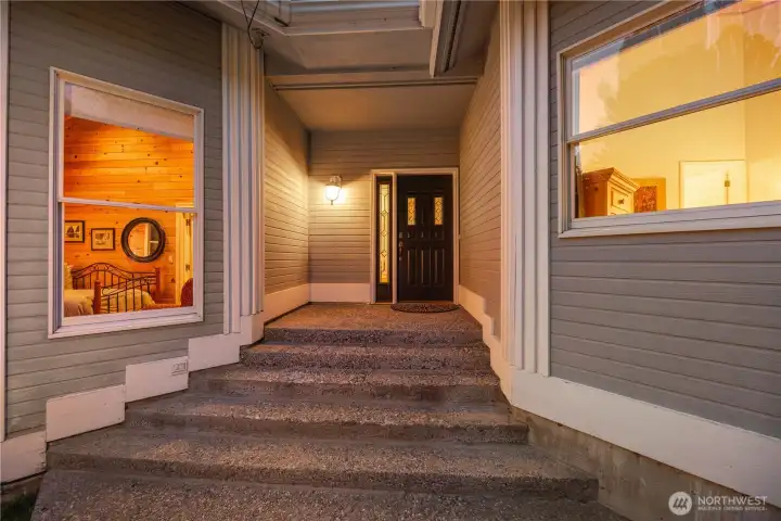 Covered entryway with wide steps leading to a recessed front door, framed by clean architectural lines and large windows. Warm interior lighting creates an inviting glow, highlighting the modern design and sheltered approach.