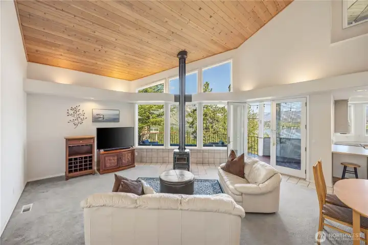 Bright living room with vaulted wood tongue and groove ceiling, large picture windows, and abundant natural light. A central fireplace anchors the space, complemented by neutral finishes and an open, airy layout.