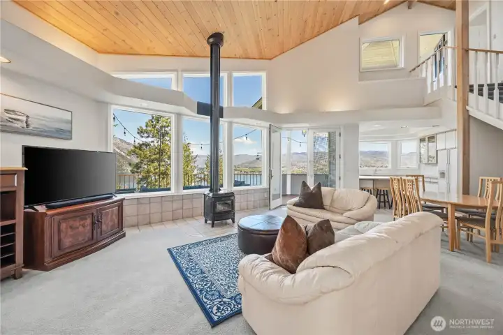 Open living area with vaulted ceiling, expansive windows, and a central freestanding stove. The space features a comfortable seating area, and an airy layout that flows into the adjacent kitchen.