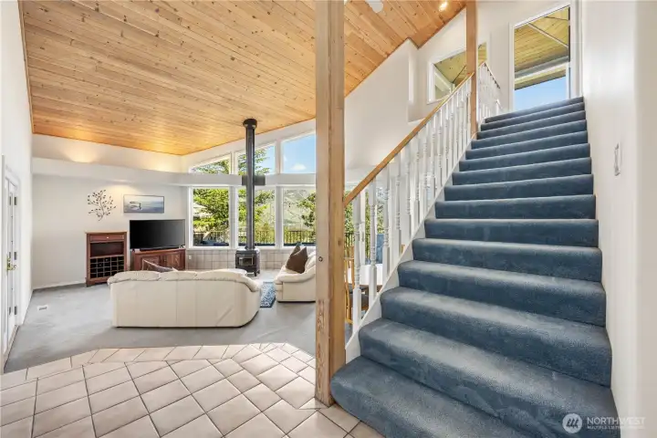 Open living area with vaulted wood-paneled ceiling, large windows, and abundant natural light. A staircase with wood railing leads to the upper level, complementing the airy layout and connecting the spaces.