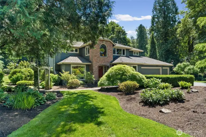The exterior features a striking brick accent wall with an arched window, anchoring the home's classic architecture amid mature trees and professionally landscaped grounds. A meandering paver pathway invites you through vibrant garden beds, showcasing the kind of curb appeal that turns heads from the street.