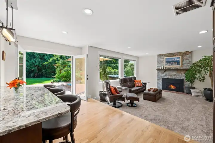 Gather around the brick gas fireplace in this open-concept great room, where hardwood flows seamlessly into a carpeted seating area framed by Marvin casement windows and French doors to the backyard. Hunter Douglas Silhouette shades soften the natural light, while the granite-topped island peeks in from the kitchen for effortless indoor-outdoor living. Notice the lush garden views just beyond the glass ~ your private backdrop for relaxing evenings by the fire.