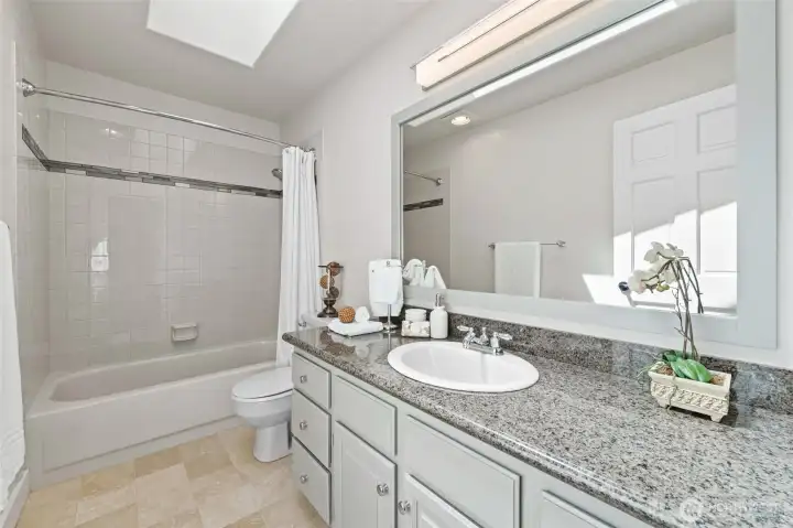 The main hall bathroom features a granite vanity, a tiled tub/shower combination with decorative accent tile, and a large mirror that reflects natural light from the skylight above. Notice the rich tile flooring — a timeless, low-maintenance finish that stands up beautifully to everyday life.
