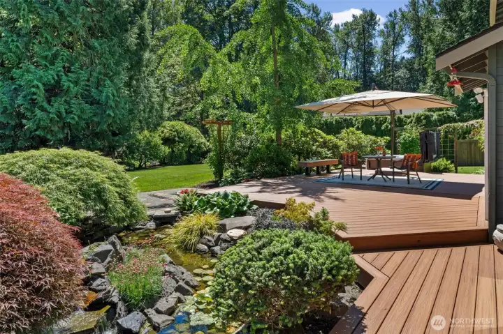 The backyard features an expansive, beautifully kept Trex brand deck overlooking a water feature with a cascading waterfall, surrounded by vibrant Japanese maples and lush greenery. Don't miss the al fresco dining setup beneath the umbrella, ideal for entertaining guests in a garden setting.
