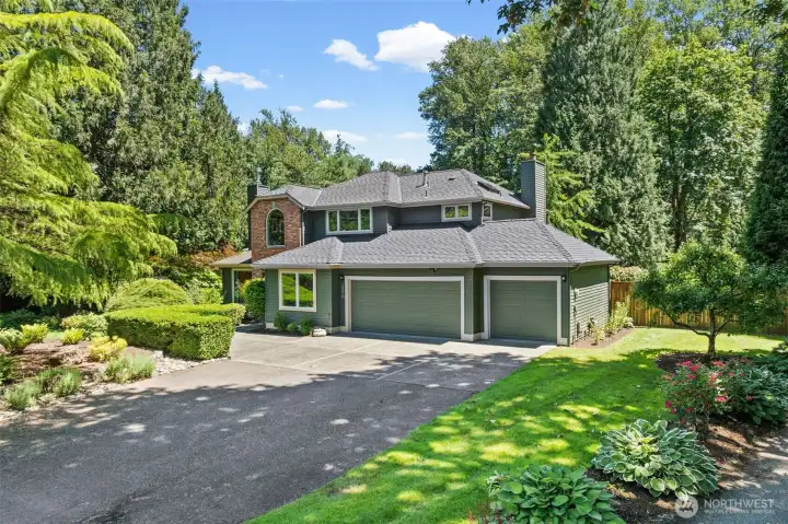 The exterior features a freshly maintained three-car garage and an expansive driveway, framed by mature evergreens and manicured hedges for striking curb appeal. Layers of vibrant landscaping, including blooming roses and hostas, create a polished first impression that signals true pride of ownership.
