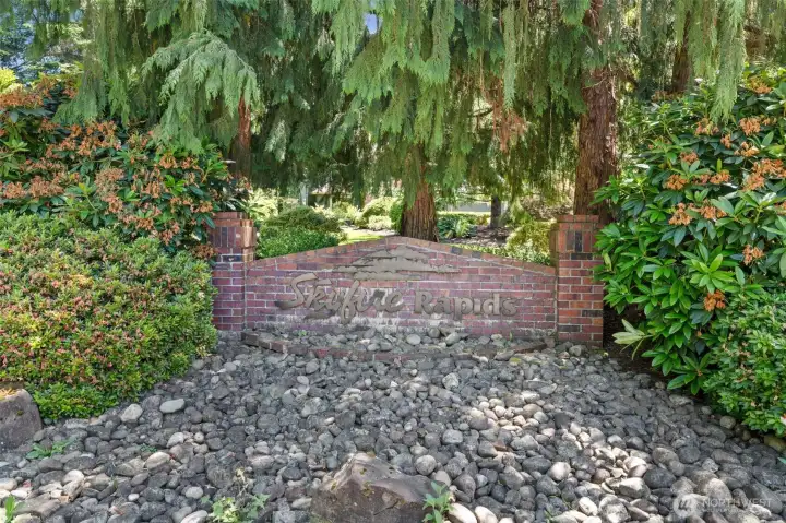 A striking brick monument framed by towering cedars and blooming rhododendrons marks the entrance to the sought-after Skyfire Rapids community, known for its well-maintained HOA and active neighborhood events.