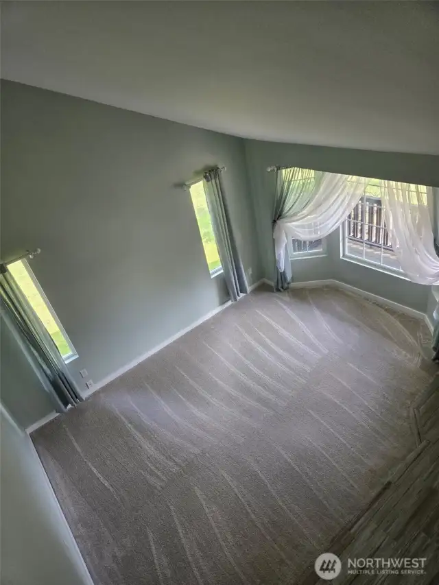 Aerial view of the sun-filled living room highlights soaring vaulted ceilings, tall windows, and fresh carpet. This elegant entry space sets the tone for warmth and brightness throughout the home.