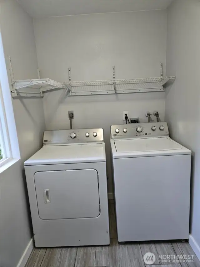 Convenient main-level laundry space features a side-by-side washer and dryer set, updated tile flooring, and upper wire shelving for added storage. A window adds natural light to brighten laundry days.