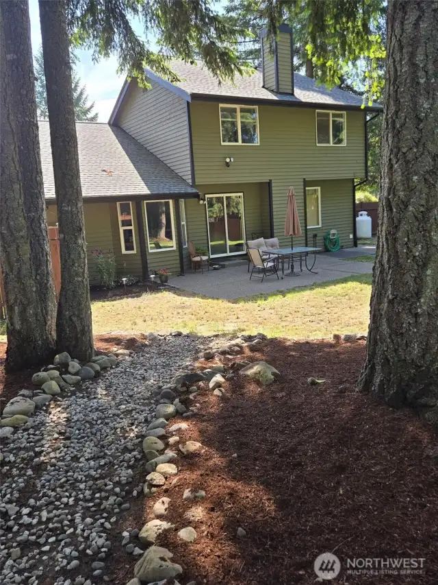 A natural woodland path leads from towering evergreens to the home's back patio, offering a seamless transition between nature and outdoor living. This backyard retreat blends tranquility with usability.