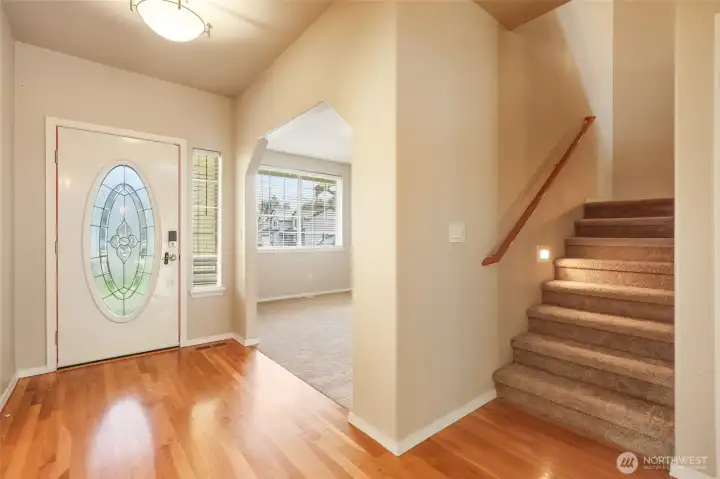 Welcome home to warmth, light, and timeless detail. Gleaming hardwood floors and a decorative glass entry door create a bright and inviting first impression, while an elegant architectural opening reveals a versatile flex room just beyond. The staircase, accented with a warm wood railing and integrated step lighting, adds both safety and sophistication — a graceful transition that sets the tone for comfort and refined living throughout.