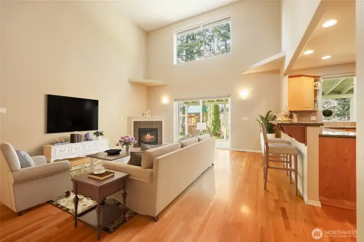 The dramatic great room is defined by soaring cathedral ceilings and expansive windows that flood the space with natural light. Rich hardwood floors flow seamlessly throughout, while a cozy gas fireplace creates a warm focal point for gathering. Open to the kitchen and inviting breakfast bar, this thoughtfully designed layout encourages effortless entertaining and everyday connection, with sliding doors extending living outdoors to the covered patio and greenbelt beyond.