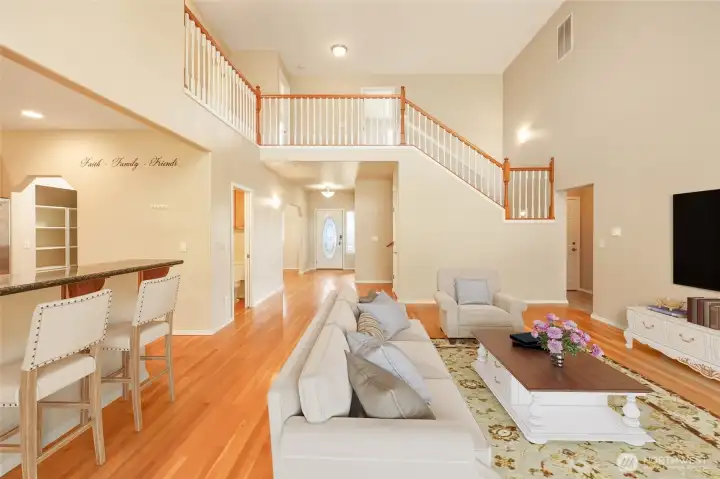 Soaring ceilings and an open second-story overlook define this dramatic yet inviting great room. Hardwood floors flow seamlessly across the main level, connecting the entry, living area, and kitchen with breakfast bar seating. A staircase accented with warm wood railing enhances the architectural character, while the expansive layout delivers an airy, light-filled setting ideal for gathering and entertaining.
