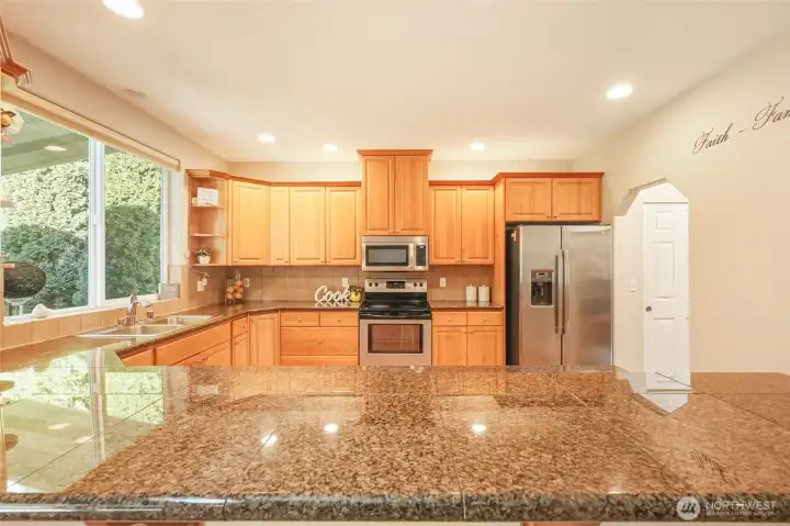 The chef’s kitchen showcases rich cabinetry, gleaming granite countertops, and stainless steel appliances, all thoughtfully arranged for both beauty and function. Recessed lighting enhances the warm tones, while the expansive breakfast bar provides generous seating for casual dining and conversation. A window over the sink brings in natural light and views of greenery, and the adjacent closet pantry ensures organized storage for everyday convenience.