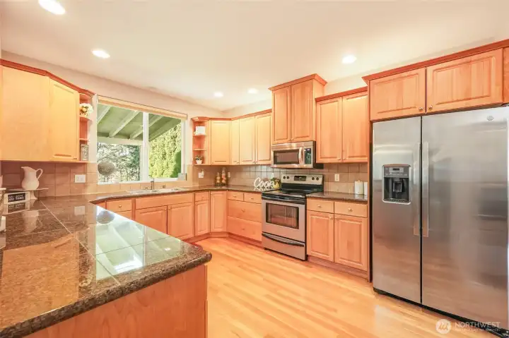 The chef’s kitchen is beautifully appointed with custom-built wood cabinetry accented by crown molding, gleaming granite countertops, stainless steel appliances, and recessed lighting that enhances both warmth and functionality. An expansive L-shaped layout provides generous prep space, while the large window over the sink frames views of the covered patio and greenery beyond. Hardwood floors continue seamlessly throughout, and the spacious breakfast bar offers ideal seating for casual dining and effortless entertaining.
