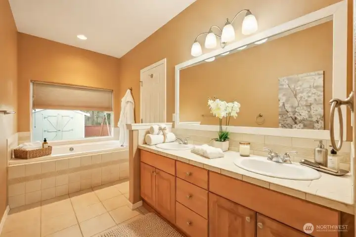 The spacious primary bath offers a spa-inspired retreat with a double vanity, abundant cabinetry, and generous tile flooring. A deep soaking tub sits beneath a sunlit window, creating the perfect place to unwind. Warm finishes and thoughtful layout combine comfort, functionality, and timeless appeal.