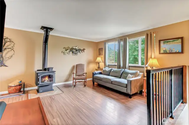 Bright and welcoming living room with large windows, outdoor views and a cozy wood-burning stove. Stylish laminate flooring and warm tones create a comfortable, inviting space.