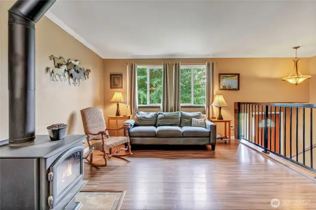 Cozy and light-filled living area featuring a wood-burning stove, scenic outdoor views, and warm laminate flooring—perfect for relaxing or gathering with guests.