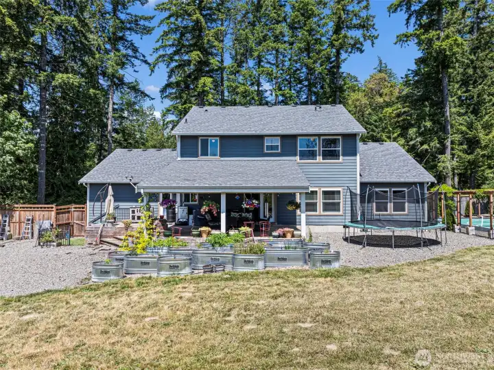 Facing back of house with raised gardens, covered porch, trampoline, sport court, chicken coop & firepit.