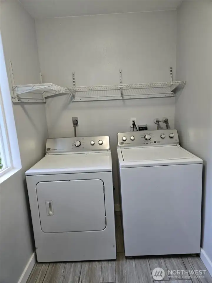 Convenient main-level laundry space features a side-by-side washer and dryer set, updated tile flooring, and upper wire shelving for added storage. A window adds natural light to brighten laundry days.