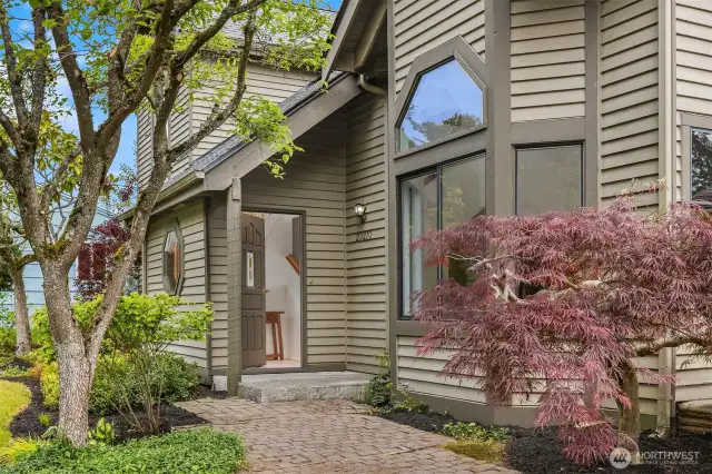 Inviting covered front entry framed by trees and brick path.