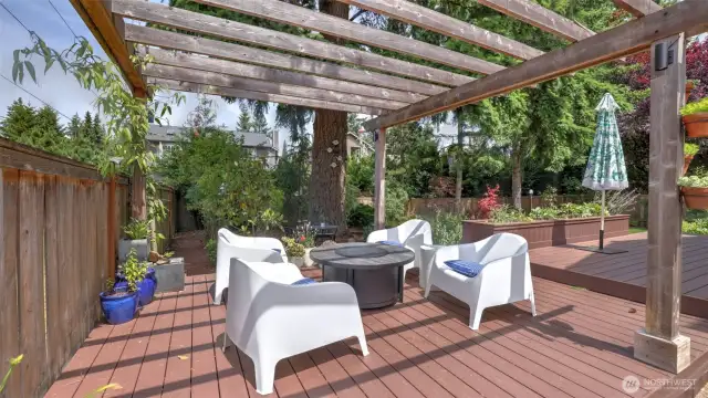 This spacious wooden deck showcases a charming two-tiered pergola, offering room for cozy seating and a fire pit. Surrounded by hanging greenery and perfect for an array of potted plants, it’s a serene outdoor retreat ready to be personalized