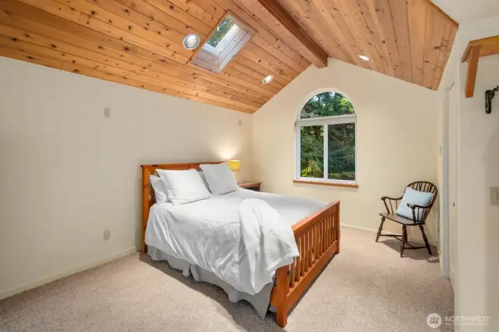 Cozy guest bedroom featuring vaulted wood ceilings, skylight, and arched window that fills the space with natural light — a warm and inviting retreat surrounded by nature.
