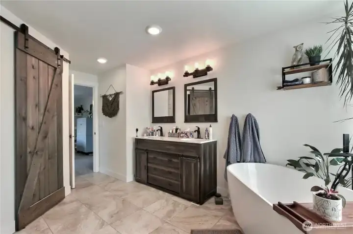 This photo showcases a master bathroom that incorporates a modern and stylish design element – a barn door leading to the bathing area, specifically featuring a free standing soaker tub. This combination of rustic barn door aesthetics with the contemporary look of a glass tub creates a unique and appealing space.