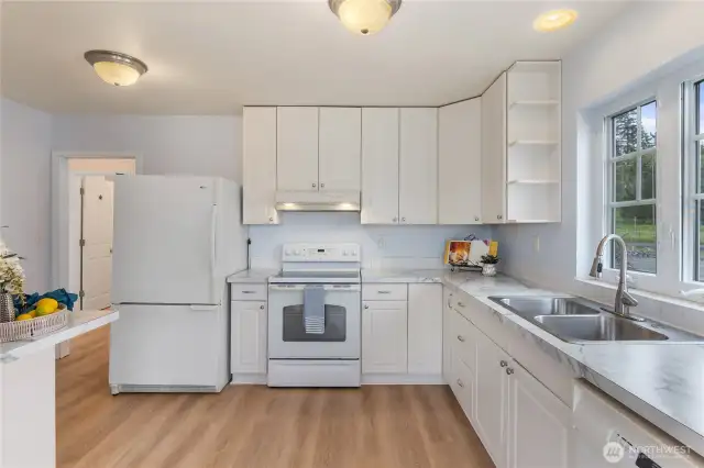 The kitchen is a welcoming space filled with natural light pouring in through generous windows. The overall feel is fresh, open, and homey, with an emphasis on comfort and simplicity.