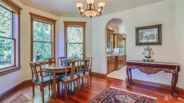 This inviting dining area offers large windows with classic wood trim, the room is bathed in natural light and showcases tranquil views of lush greenery. The open layout seamlessly connects the dining area to the adjacent kitchen through an elegant archway, enhancing the home's flow and functionality.