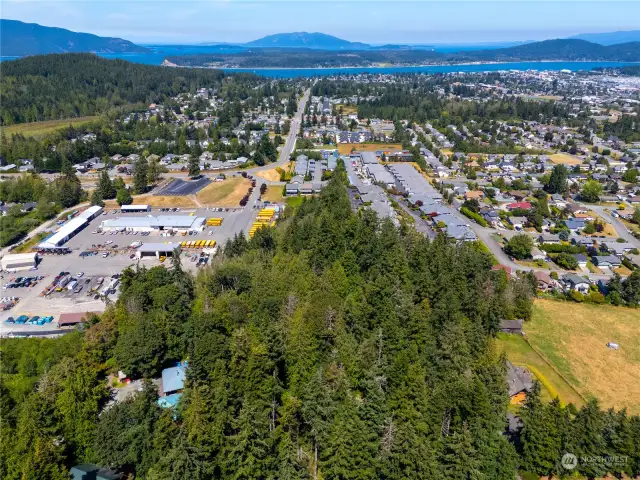 Expansive aerial view showcasing a blend of residential neighborhoods, industrial space, and lush forest, with stunning water and island views in the distance.