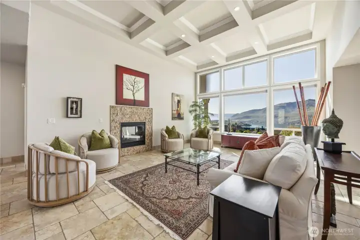 Light-filled living room featuring soaring coffered ceilings, expansive picture windows, and captivating Lake Chelan and mountain views. A cozy gas fireplace anchors the space, adding warmth and ambiance, while the thoughtful layout creates an inviting setting for both everyday living and entertaining.