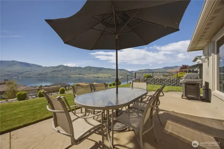 Inviting outdoor dining area on the expansive patio, complete with shade umbrella and seating for gatherings, all set against sweeping panoramic views of Lake Chelan and the surrounding mountains. The perfect setting for alfresco dining, morning coffee, or evening sunsets.