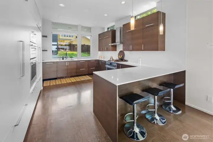 Eating bar facing the open kitchen, featuring walnut and matte white cabinetry lined with quartz countertops and a full suite of appliances, including double dishwashers and double ovens