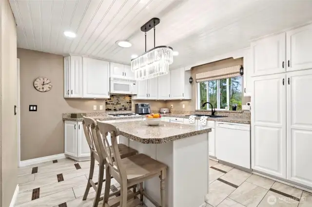 The revamped kitchen includes an island with seating, ceramic tile flooring, white cabinetry with plenty of storage, granite counter tops, and a gas cooktop.