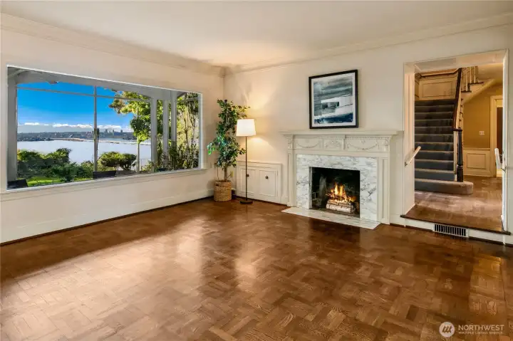 Framed by a panoramic picture window, the living room captures breathtaking views of Lake Washington and the Seattle skyline beyond, bringing the serenity of nature into the heart of the home. A classic fireplace adds warmth and ambiance, while original architectural details—like the parquet flooring and crown molding—speak to the timeless quality of the residence. Just beyond the archway, the central staircase hints at the rich story that unfolds across the home’s multiple levels.