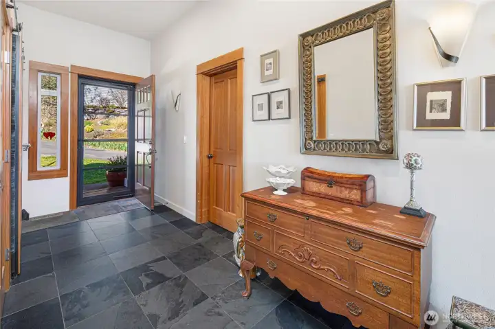 The elegant entry foyer sets a sophisticated tone with rich slate tile flooring, warm wood-trimmed doors, and tall ceilings. Glass sidelights bathe the space in natural light while providing a gracious welcome. Secondary bedroom door in view.