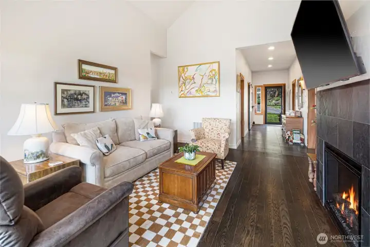 Inviting main-level living room looking down the hall to the entry, showcases rich dark hardwood and slate floors, a classic tile-surround electric fireplace, and an abundance of natural light. Hallway leads to bedrooms on the right and garage, laundry room and guest bathroom to the left.