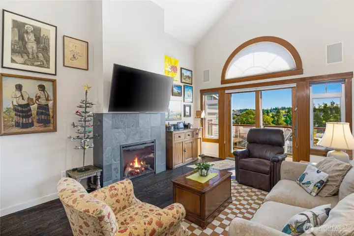 Light-filled living area with vaulted ceilings, a cozy tile-surround electric fireplace, mounted flat-screen TV, and an iconic arched window overlooking Friday Harbor, creating a warm and sophisticated gathering space.