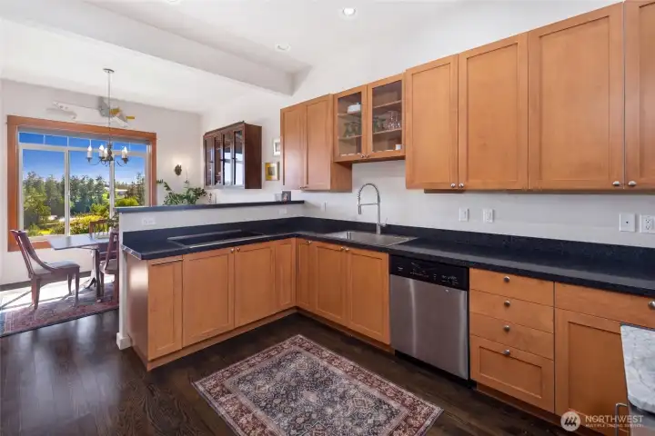 Well-appointed kitchen features warm honey-toned cabinetry, sleek dark granite countertops, stainless steel dishwasher, and a pass-through to the dining area, all bathed in natural light from the dining room window.
