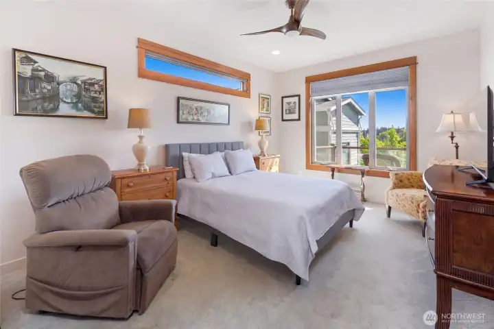 This well-appointed primary bedroom features plush carpet, warm wood window trim, and a large window with sweeping views of the surrounding neighborhood and treetops. A ceiling fan and ample natural light make this a comfortable and inviting retreat.