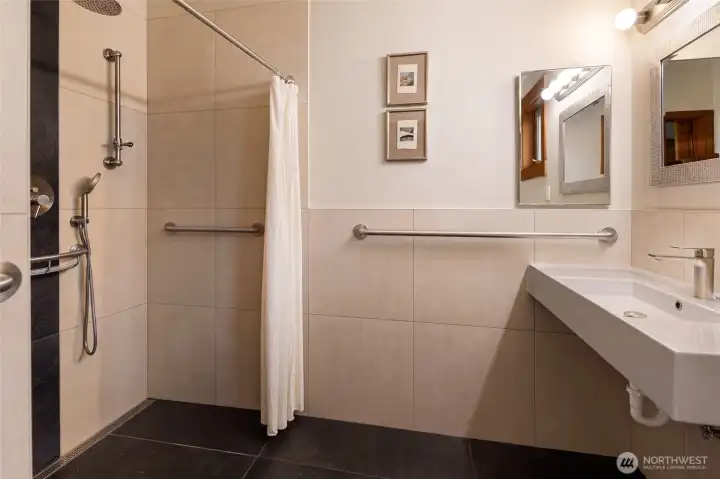 The thoughtfully designed accessible bathroom boasts a spacious roll-in shower, with premium grab bars, a sleek wall-mounted sink, and elegant large-format heated tile throughout for a clean, contemporary aesthetic. This space offers both sophisticated style and inclusive functionality for all residents and guests.